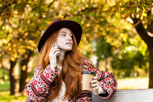Concentrated young student redhead girl in autumn park talking by mobile phone drinking coffee. - Valokuva, kuva