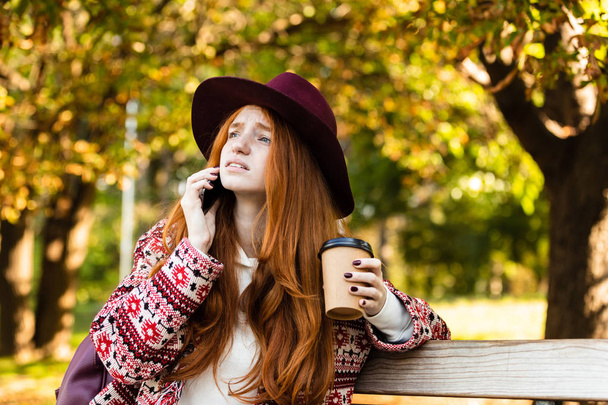 Sad young student redhead girl in autumn park talking by mobile phone drinking coffee. - Foto, Imagem