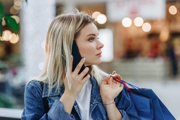 Closeup profile of beautiful blonde young woman in shopping mall talking on the phone. Beauty woman in a denim jacket with shopping bags standing in shopping mall. Copy space on the right side - Fotoğraf, Görsel