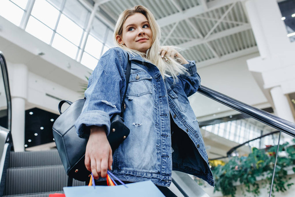 Young woman with shopping bags in shopping mall on escalator and looking aside. Young shopaholic in a denim jacket. Sales, shop, retail, consumer concept. Copy space on the left or right side - Foto, Imagem