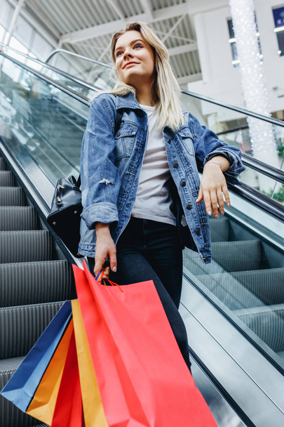 Vertical photo of young blonde woman with colorful shopping bags in shopping mall on escalator. Young shopaholic in a denim jacket. Sales, shop, retail, consumer concept - Photo, Image
