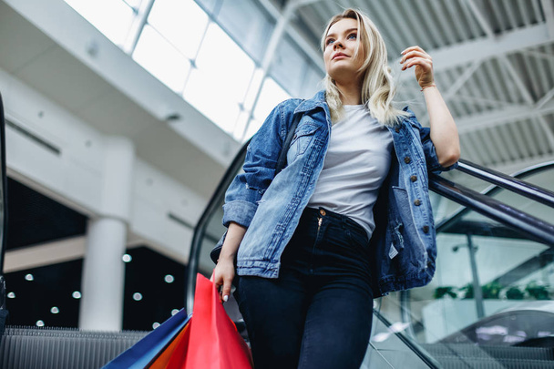 Portrait of young woman with shopping bags in shopping mall on escalator and looking aside. Young shopaholic in a denim jacket. Sales, shop, retail, consumer concept. Copy space on the left side - Foto, Imagen