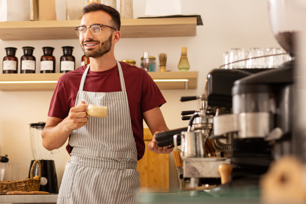 Barista alegre em pé perto da máquina de café com xícara de café
 - Foto, Imagem