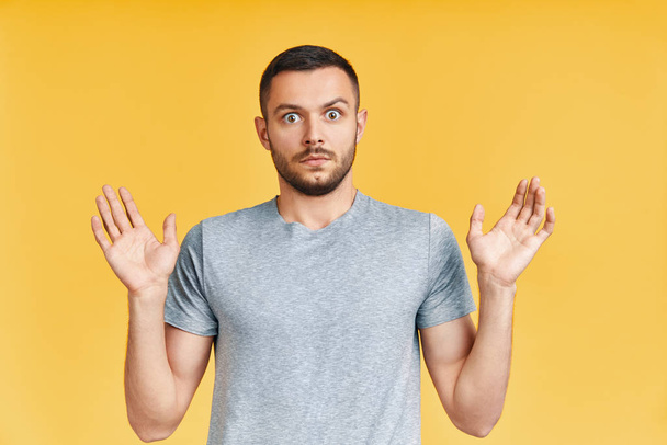 Shocked young man with arms up looking amazed in camera over yellow background. Emotions concept                 - Fotoğraf, Görsel