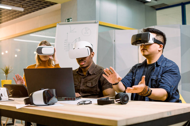 Młoda Grupa wieloetnicznych ludzi bawią się z nowymi technologiami VR Headset. Kaukaski dziewczyna, afrykańskich i chińskich mężczyzn siedzi na biurku i próbuje Gogle VR w nowoczesnym biurze - Zdjęcie, obraz