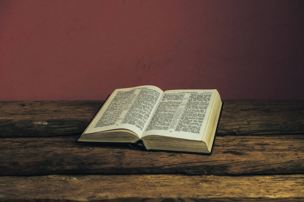 Piękne otwarte Pismo Święte na Starym dębowym drewnianym stole i ciemnym czerwonym tle ściany. Koncepcja religijna. - Zdjęcie, obraz