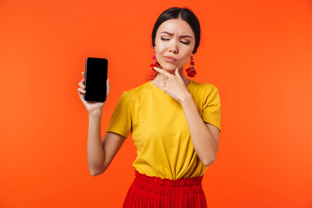 Serious young woman posing isolated over orange wall background showing display of mobile phone. - Photo, Image