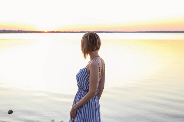 piękna dziewczyna patrzy na zachód słońca w pobliżu rzeki. Piękny czerwony zachód słońca w lecie - Zdjęcie, obraz