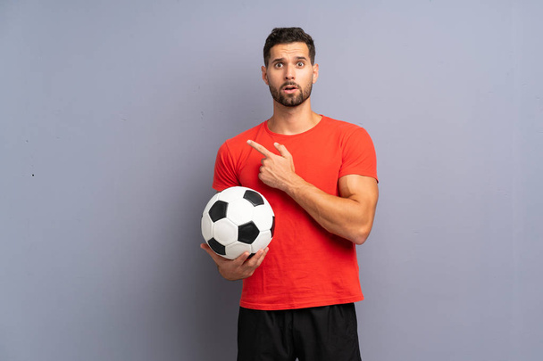 Beau jeune joueur de football homme sur mur blanc isolé surpris et pointant côté
 - Photo, image