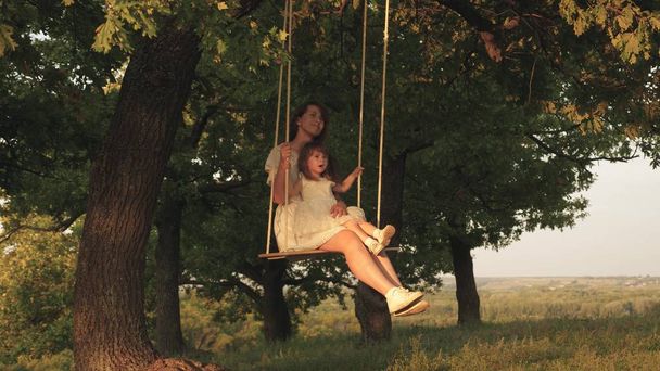 mother and baby ride on a rope swing on an oak branch in forest. Mom shakes her daughter on swing under a tree in sun. Girl laughs, rejoices. Family fun in park, in nature. warm summer day. - Foto, Imagen
