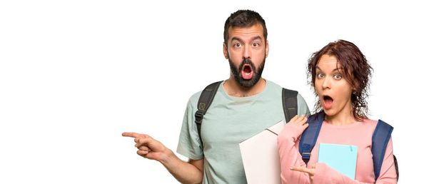 Two students with backpacks and books pointing finger to the side with a surprised face on isolated white background - Photo, Image