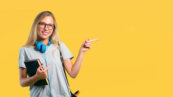 Student girl with glasses pointing finger to the side and presenting a product while smiling in a confident pose on yellow background - Фото, изображение
