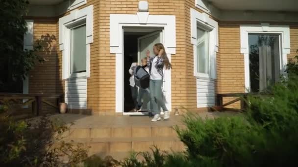 Little cute boy and girl with backpacks leaving the house and going to school. Smiling mother waving a hand. Happy loving family concept - Materiaali, video