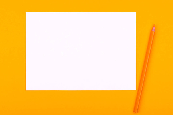 Ένα λευκό φύλλο χαρτί για σχέδια, κείμενα και νότες δίπλα σε ένα μολύβι βρίσκεται σε πορτοκαλί φόντο - Φωτογραφία, εικόνα