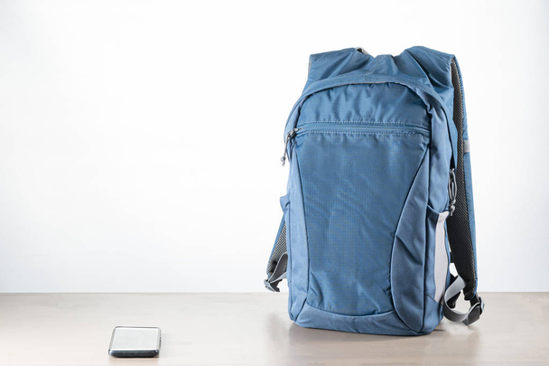 Niebieski plecak do szkoły na brązowym drewnianym stole obok telefonu komórkowego z białą ścianą. Powrót do szkoły - Zdjęcie, obraz