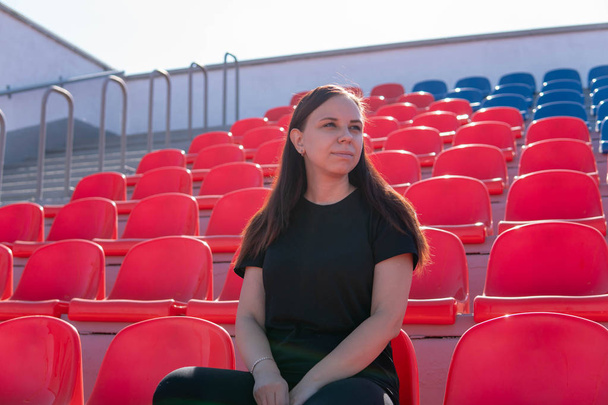 Eine junge Frau in schwarzer Kleidung mit langen Haaren sitzt allein auf der Tribüne des Stadions und verfolgt ein Sportspiel unter sengender Sonne.. - Foto, Bild