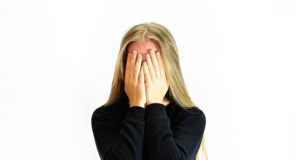 A beautiful girl with long blonde hair covers her face with a ring on her right hand wearing a black sweater located in the center of the photo, waist-high photo on a white background - Foto, immagini