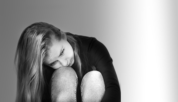 Sad beautiful girl with long blonde hair dressed in jeans and a black sweater rested her head on her bent knees, photo in black and white format on a white background - Photo, Image