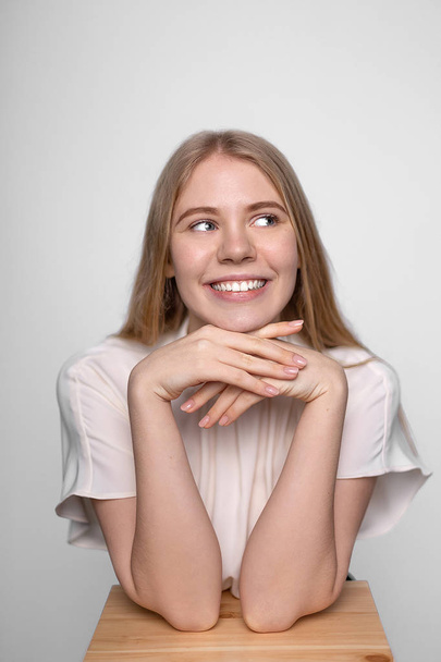 Young smiling blonde girl puts her head on hands, rests on stool, playfully looking aside. Portrait on white background - Valokuva, kuva