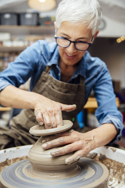 彼女のワークショップに座っている間、陶器の車輪に取り組む格好良い先輩の女性陶芸家 - 写真・画像