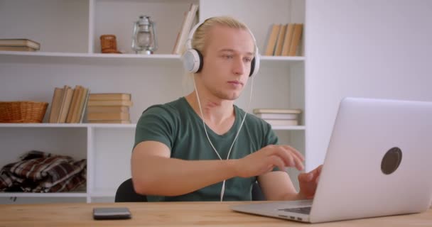 Closeup portrait of young handsome blonde hipster businessman in headphones using laptop being relaxed indoors in office - Imágenes, Vídeo