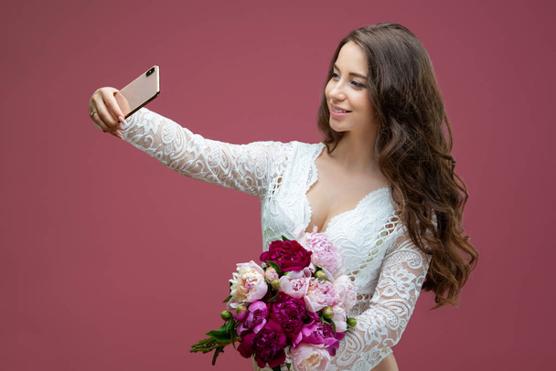 Beautiful pregnant woman in lace white lingerie body suit makes a selfie with a bouquet of flowers peony - Foto, Imagem