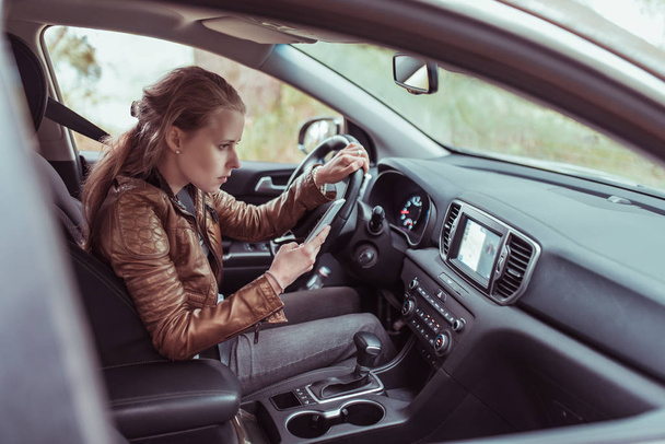 woman driving car, selects navigation on phone s display screen, turns voice assistant, searches for route to Internet, checks map with phone navigation in car. Leather jacket summer autumn on trip. - Фото, изображение