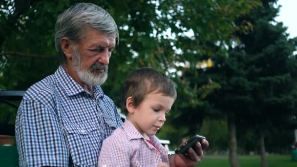 Abuelo mayor y nieto están sentados en un banco en el parque y jugando en un teléfono inteligente
 - Imágenes, Vídeo