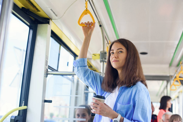 Beautiful young woman drinks delicious coffee in city bus or tram. Concept of public transport. - Photo, Image