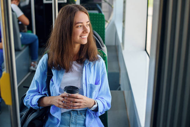Όμορφο κορίτσι με φλιτζάνι νόστιμο καφέ βόλτες με το Πανεπιστήμιο στις δημόσιες συγκοινωνίες. - Φωτογραφία, εικόνα