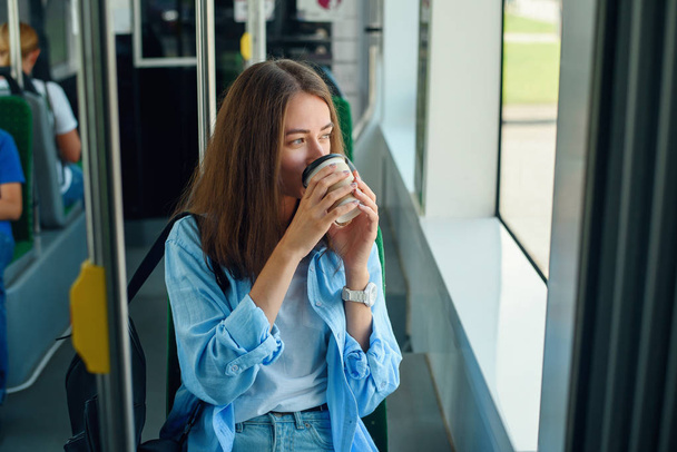 Beautiful young woman drinks delicious coffee in city bus or tram. Concept of public transport. - Φωτογραφία, εικόνα
