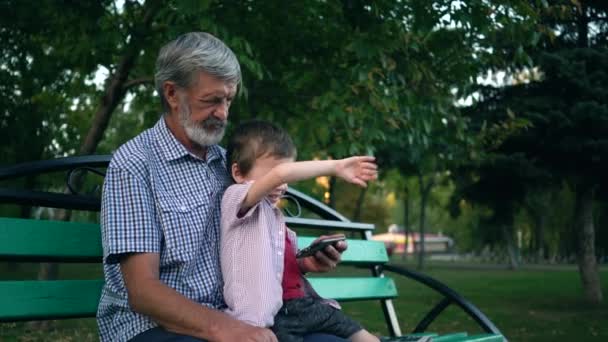 Grand-père aîné et petit-fils sont assis sur un banc dans le parc et jouent sur un smartphone
 - Séquence, vidéo