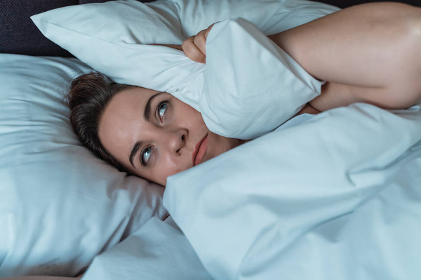 Молодая женщина в стрессе не может спать из-за шума соседей сверху, раздраженная и напряженная покрывает голову и уши подушкой
 - Фото, изображение