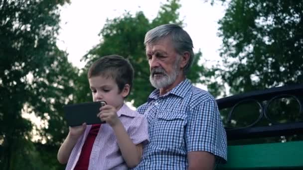 Vanhempi isoisä ja pojanpoika istuvat penkillä puistossa ja pelaavat älypuhelimella
 - Materiaali, video
