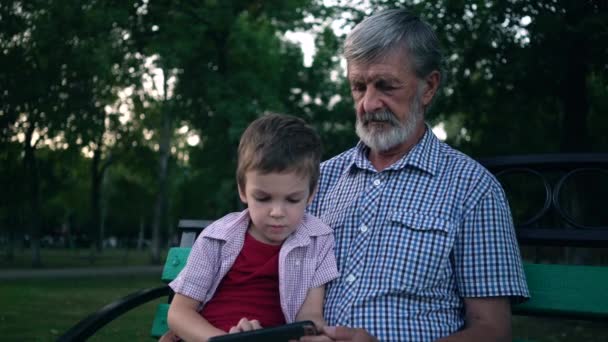 Avô e neto sênior estão sentados em um banco no parque e jogando em um smartphone
 - Filmagem, Vídeo