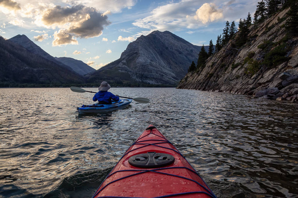 Adventurous Man Kayaking in Glacier Lake surrounded by the beautiful Canadian Rocky Mountains during a cloudy summer sunset. Taken in Upper Waterton Lake, Alberta, Canada. - Zdjęcie, obraz