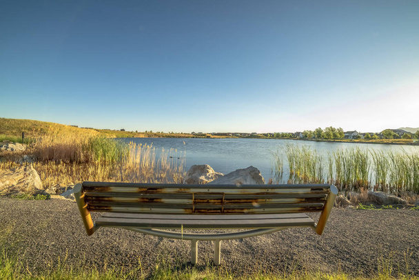 Visão traseira de um banco enferrujado de frente para um lago brilhante e céu azul claro em um dia ensolarado
 - Foto, Imagem