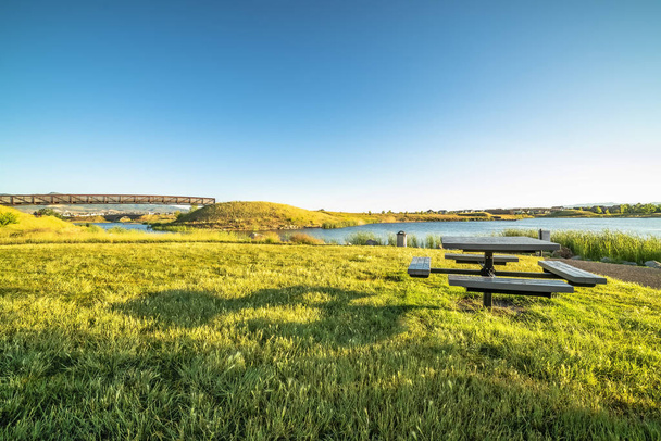 Площадка для пикника на травянистой местности с видом на озеро под ясным голубым небом
 - Фото, изображение