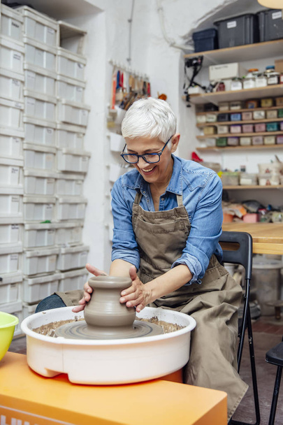 Onun atölyesinde otururken çömlek tekerleği üzerinde çalışan iyi görünümlü yaşlı kadın çömlekçi - Fotoğraf, Görsel