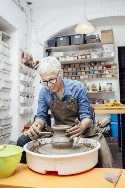 Good looking senior female potter working on pottery wheel while sitting  in her workshop - Photo, Image