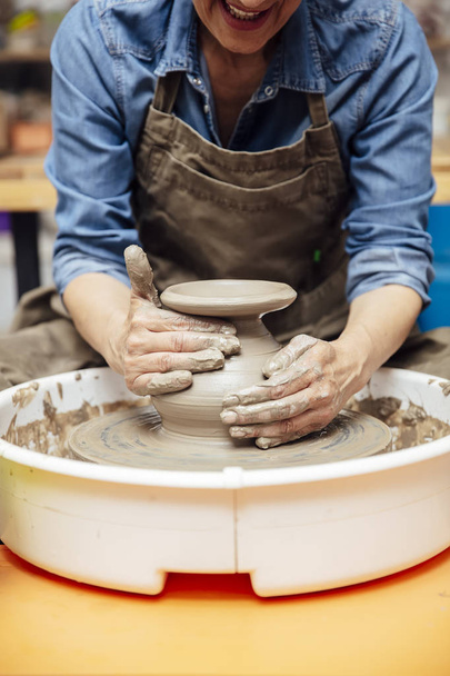 Alfarera senior de buen aspecto trabajando en rueda de cerámica mientras está sentada en su taller
 - Foto, Imagen