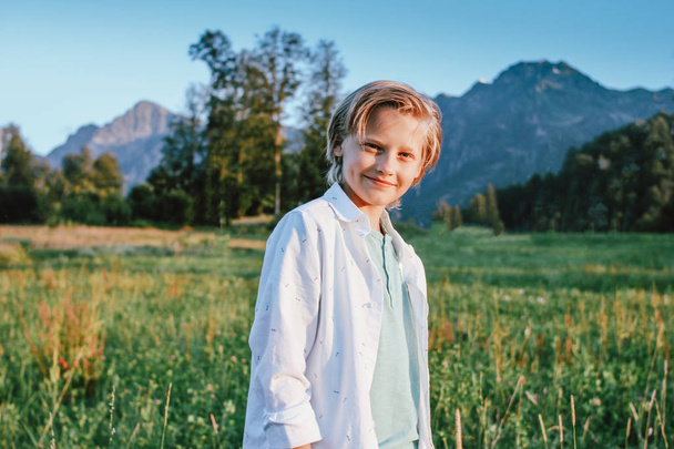 Rubia sonriente chico mirando a la cámara en el fondo de la maravillosa vista de la pradera verde y las montañas, familia viaje aventura estilo de vida
 - Foto, Imagen