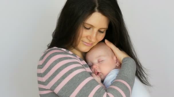長い黒髪の若い美しい母親は、スタジオで白い背景に2ヶ月の新生児の赤ちゃんを保持しています - 映像、動画