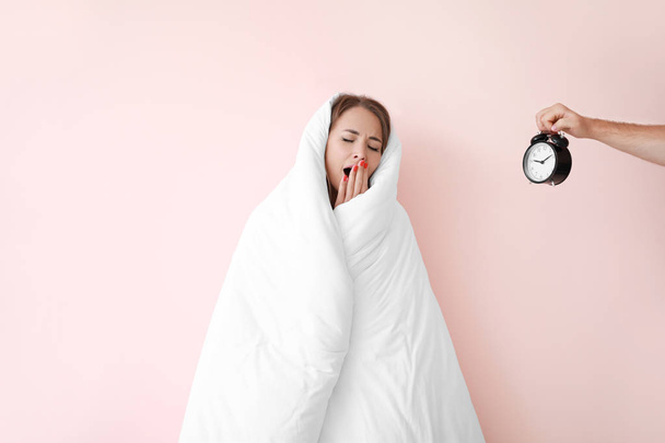 Sleepy young woman wrapped in blanket and male hand with alarm clock on color background - Photo, Image