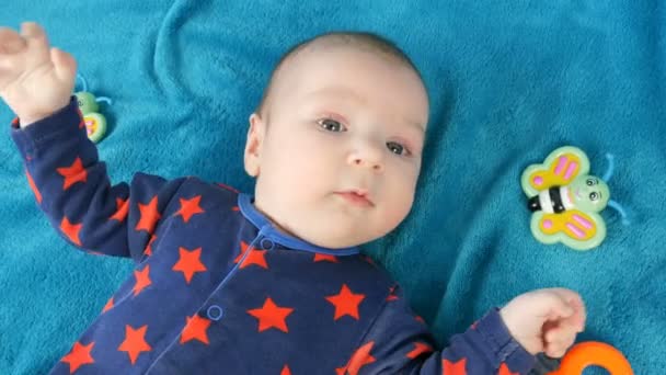 Porträt eines lustigen kleinen Neugeborenen von zwei Monaten, das auf einer blauen Bettdecke liegt, umgeben von Babyrasseln - Filmmaterial, Video