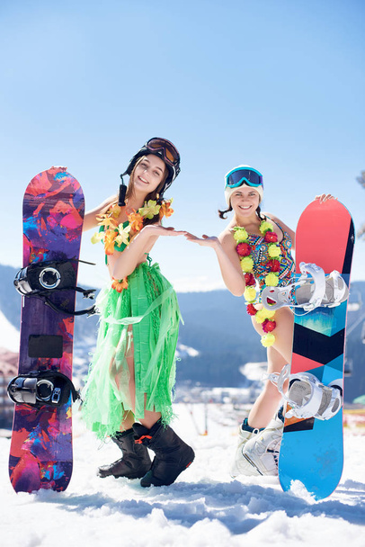Dos atractivas snowboarders femeninas en traje de baño, sujetador, falda y decoración floral con tablas de snowboard sobre fondo de cielo azul y montañas de invierno
 - Foto, Imagen