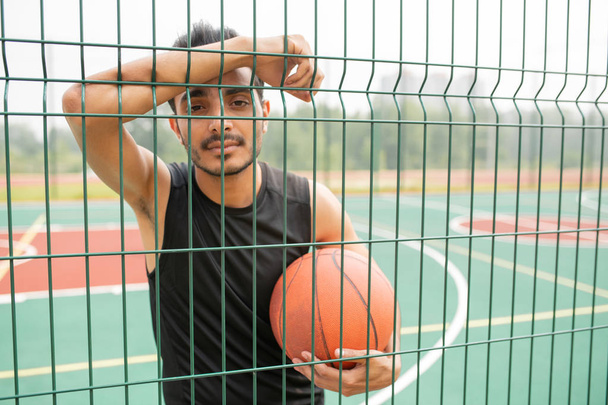Serieuze mannelijke basketbalspeler met bal staande tegen het net terwijl je naar je kijkt bij pauze op speelplaats - Foto, afbeelding