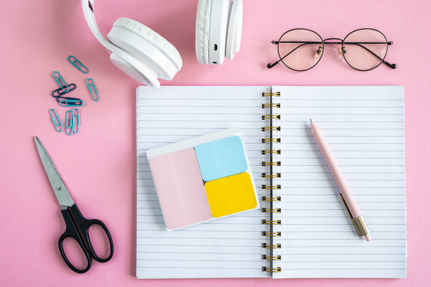 Open notebook, erasers, pen, clips, scissors, eyeglasses and headphones on pink background - Foto, Imagen