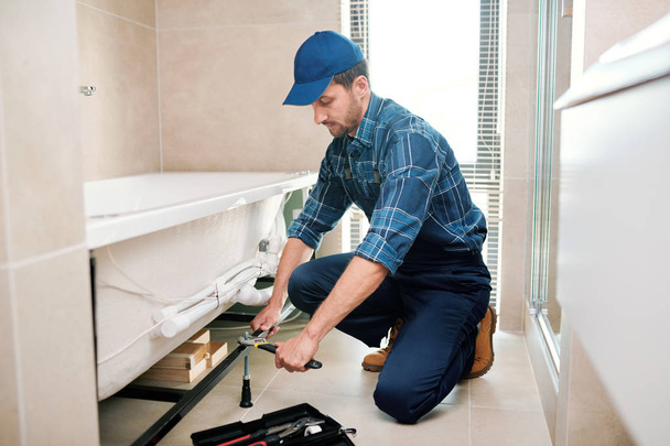 Jonge loodgieter of technicus in werkkleding voorbereiding detail voor badkuip installatie terwijl zittend op squats - Foto, afbeelding