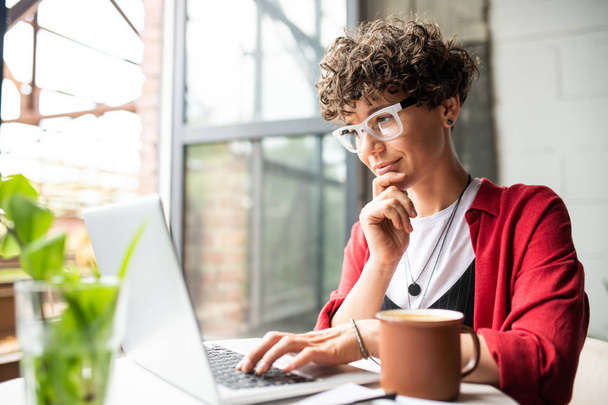 geschäftige junge elegante Frau mit Brille, die auf das Laptop-Display blickt, während sie während der Arbeit die Tasten der Tastatur drückt - Foto, Bild
